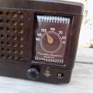 Puede incluir: Una radio Emerson marr&oacute;n vintage con un dial que muestra n&uacute;meros del 55 al 160. La radio tiene una rejilla de altavoz con m&uacute;ltiples agujeros y un mando de sintonizaci&oacute;n. El texto "Emerson Radio and Phonograph Corp New York USA" es visible.