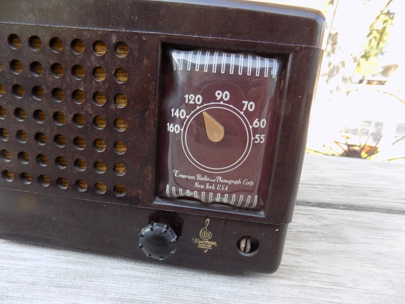 Puede incluir: Una radio Emerson marr&oacute;n vintage con un dial que muestra n&uacute;meros del 55 al 160. La radio tiene una rejilla de altavoz con m&uacute;ltiples agujeros y un mando de sintonizaci&oacute;n. El texto "Emerson Radio and Phonograph Corp New York USA" es visible.