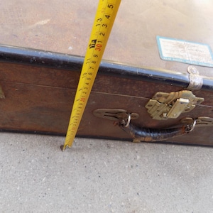 May include: A vintage brown suitcase with a black trim, metal clasps, and a leather handle. A yellow tape measure shows a measurement of approximately 12 inches. The suitcase has a label on the top.