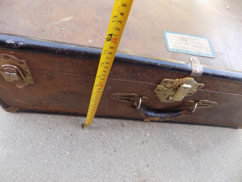 May include: A vintage brown suitcase with a black trim, metal clasps, and a leather handle. A yellow tape measure shows a measurement of approximately 12 inches. The suitcase has a label on the top.