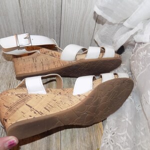K&ouml;nnte beinhalten: Wei&szlig;e Keilsandalen mit Korksohlen und einem Schnallenverschluss. Die Sandalen haben einen wei&szlig;en Lederriemen &uuml;ber dem Fu&szlig; und einen wei&szlig;en Lederriemen am Kn&ouml;chel.