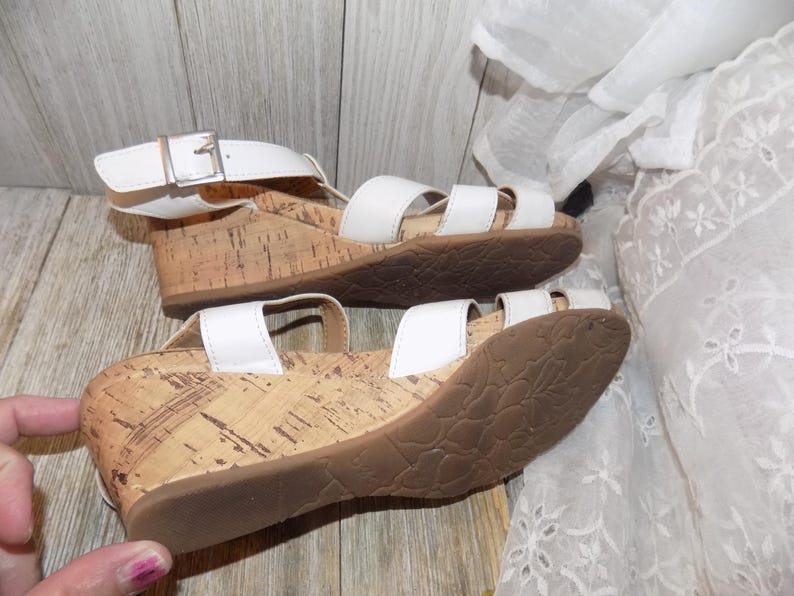 K&ouml;nnte beinhalten: Wei&szlig;e Keilsandalen mit Korksohlen und einem Schnallenverschluss. Die Sandalen haben einen wei&szlig;en Lederriemen &uuml;ber dem Fu&szlig; und einen wei&szlig;en Lederriemen am Kn&ouml;chel.