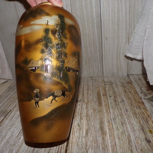 May include: A gold ceramic vase with a Japanese landscape scene painted on it. The scene includes mountains, trees, houses, and people. The vase has a glossy finish.