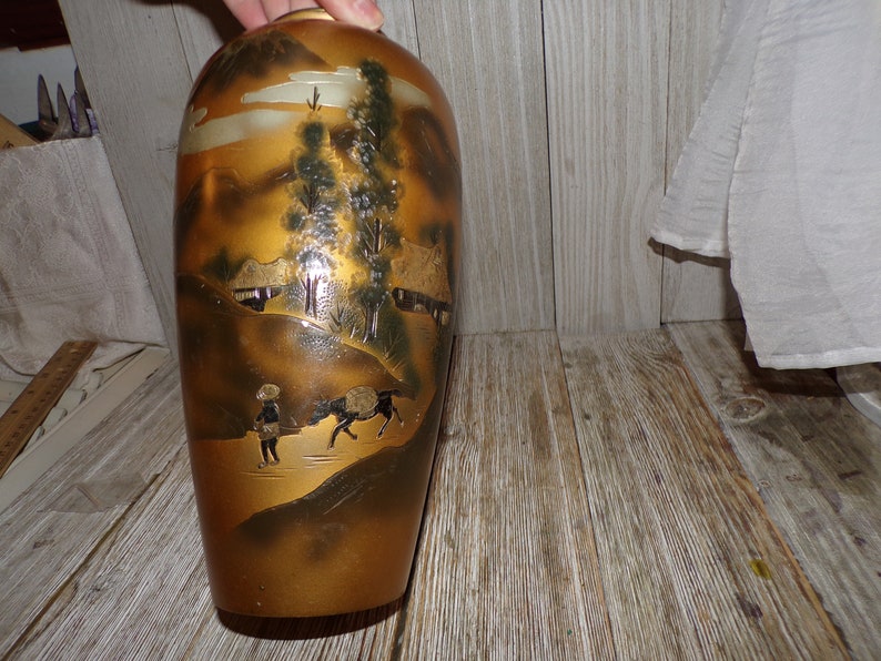 May include: A gold ceramic vase with a Japanese landscape scene painted on it. The scene includes mountains, trees, houses, and people. The vase has a glossy finish.