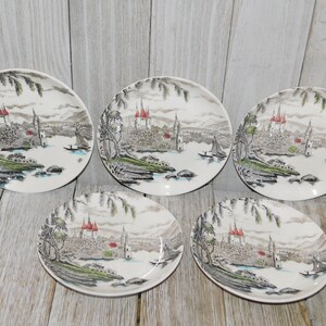 May include: Five white ceramic plates with a landscape design. Each plate features a detailed illustration of a castle with red roofs, trees, and a body of water with small boats. The plates have a vintage aesthetic.