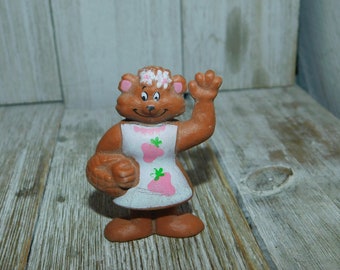May include: A brown bear figurine with a white dress adorned with pink strawberries and green leaves. The bear is waving with its right hand and holding a basket. A floral crown sits atop its head. The figurine is on a wooden surface.