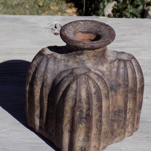 May include: A weathered, brown ceramic vase with a wide, fluted body and a circular opening. The vase has a textured surface and a dark shadow cast to the left. The overall design suggests a rustic or antique aesthetic.