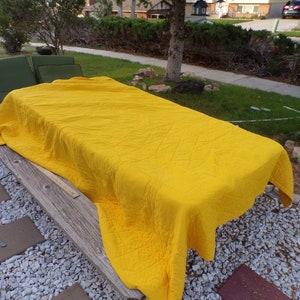 May include: A bright yellow quilt covers a wooden picnic table outdoors. The quilt has a diamond pattern and is draped over the table. A green chair and a yard with grass and a sidewalk are in the background.