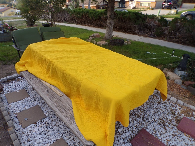 May include: A bright yellow quilt covers a wooden picnic table outdoors. The quilt has a diamond pattern and is draped over the table. A green chair and a yard with grass and a sidewalk are in the background.