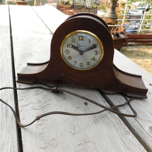 May include: A vintage wooden electric clock with a gold-colored face and black hands. The clock is plugged in and has a cord.