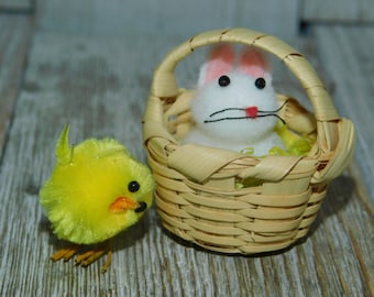 May include: A yellow chick figurine next to a small wicker basket with a white bunny figurine inside. The bunny has pink ears and black eyes.