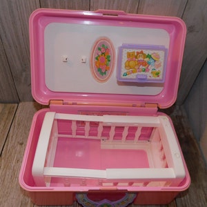 Puede incluir: Un portabeb&eacute; de pl&aacute;stico rosa con una cuna blanca en el interior. El portabeb&eacute; tiene una tapa blanca con una imagen de una cuna y una imagen de un osito de peluche con un biber&oacute;n y otros art&iacute;culos para beb&eacute;s.