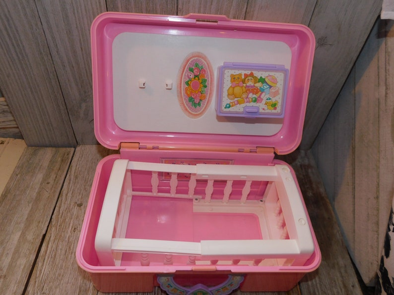 Puede incluir: Un portabeb&eacute; de pl&aacute;stico rosa con una cuna blanca en el interior. El portabeb&eacute; tiene una tapa blanca con una imagen de una cuna y una imagen de un osito de peluche con un biber&oacute;n y otros art&iacute;culos para beb&eacute;s.