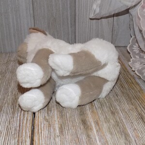 May include: A plush toy cat in a lying position, with its underside visible. The cat has white fur with brown patches on its paws and ears. The toy is on a wooden surface.