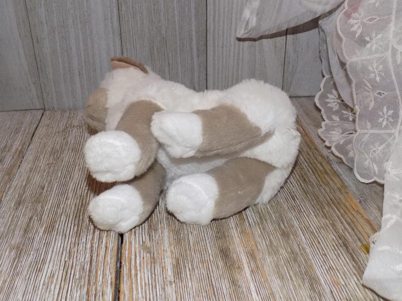 May include: A plush toy cat in a lying position, with its underside visible. The cat has white fur with brown patches on its paws and ears. The toy is on a wooden surface.