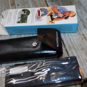 May include: A black leather case with a silver button and the word "MICRONTA" printed on it. The case is closed and is sitting on a wooden surface. There is a box in the background with a blue and white design.