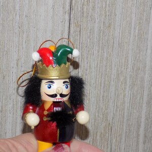 May include: A wooden nutcracker ornament with a red and black jester costume, a green and red hat, and a gold crown. The nutcracker is holding a white ball in each hand.