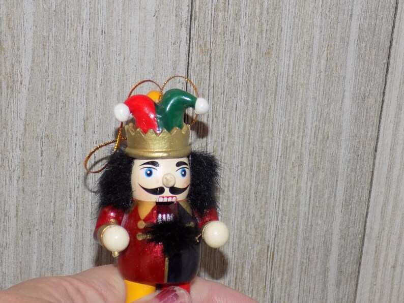 May include: A wooden nutcracker ornament with a red and black jester costume, a green and red hat, and a gold crown. The nutcracker is holding a white ball in each hand.
