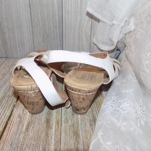 K&ouml;nnte beinhalten: Ein Paar wei&szlig;e Keilsandalen mit Korkabs&auml;tzen. Die Sandalen haben einen Riemen &uuml;ber dem Fu&szlig; und einen Kn&ouml;chelriemen.