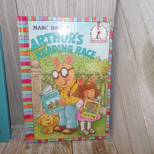 May include: A children's book titled "Arthur's Reading Race" by Marc Brown. The cover features Arthur and his sister D.W. reading books. Arthur is holding a book titled "Soccer" and D.W. is holding a book titled "Bears". The book is part of the "I Can Read It All By Myself" Beginner Books series.