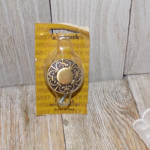May include: A gold-toned cabinet knob with a decorative, intricate design in black. The knob is packaged on a yellow card with the text "Renaissance" and "Antiqued English" printed on it.