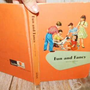 May include: An orange hardcover book titled Fun and Fancy Revised Edition. The cover features an illustration of four children interacting with a spotted dog. The spine displays the title and the publisher name Ginn. A Ginn Basic Readers logo is visible.
