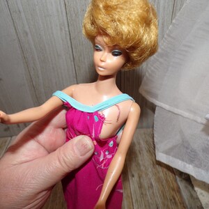 May include: A vintage Barbie doll with blonde hair wearing a pink and blue dress. The dress has a floral pattern and a blue trim. The doll's hair is styled in a short, curly bob.