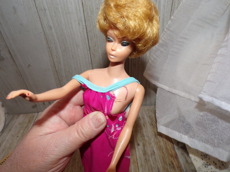 May include: A vintage Barbie doll with blonde hair wearing a pink and blue dress. The dress has a floral pattern and a blue trim. The doll's hair is styled in a short, curly bob.