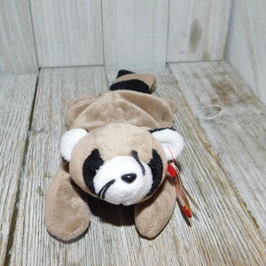 K&ouml;nnte beinhalten: Ein braunes und schwarzes Waschb&auml;r-Beanie Baby mit wei&szlig;em Gesicht und schwarzen Augen. Das Beanie Baby liegt auf der Seite mit eingerolltem Schwanz. Das Beanie Baby hat ein rotes und wei&szlig;es Etikett.