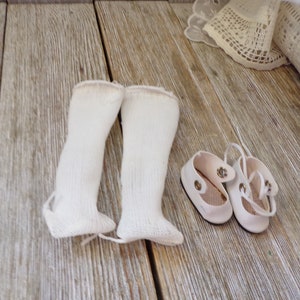 May include: Two pairs of white doll socks and two pairs of white doll shoes. The socks are knitted and the shoes have a strap.