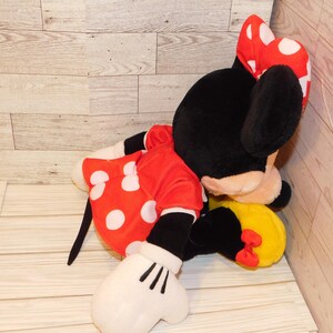 Op de afbeelding: Een pluche Minnie Mouse pop, met een zwarte kop en oren, een rode jurk met witte stippen en een bijpassende strik. De pop heeft witte handschoenen en een gele rok met een rode strik. Het speelgoed ligt op een licht houten oppervlak.