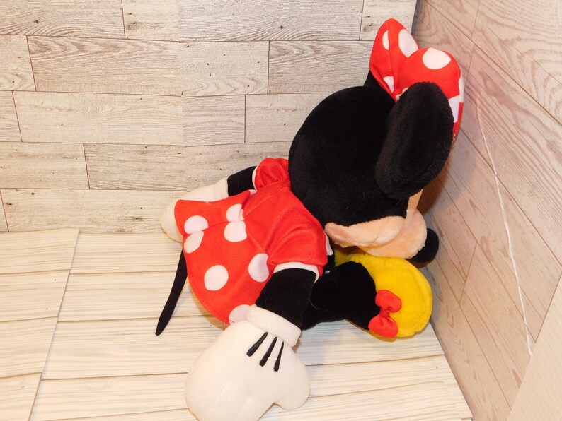 Op de afbeelding: Een pluche Minnie Mouse pop, met een zwarte kop en oren, een rode jurk met witte stippen en een bijpassende strik. De pop heeft witte handschoenen en een gele rok met een rode strik. Het speelgoed ligt op een licht houten oppervlak.