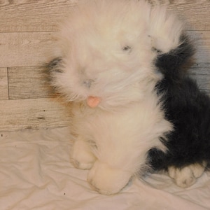 May include: A plush Old English Sheepdog stuffed animal with a fluffy white body and a dark gray tail. The toy has a pink tongue and is sitting on a white surface.