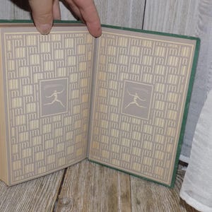 May include: A green hardcover book with a patterned design on the cover. The pattern is a series of small squares with a stylized figure in each square. The book is open to reveal the same pattern on the inside pages.