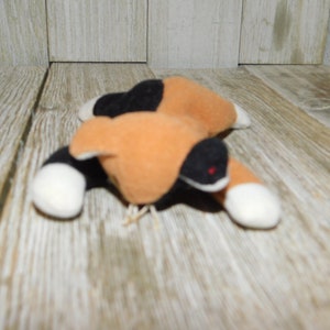 May include: A small, plush calico cat toy with black, orange, and white fur. The cat is lying on its belly with its paws outstretched.