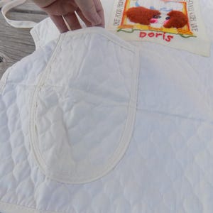Op de afbeelding: Witte gewatteerde schort met een grote zak en een geborduurde patch met de tekst "The fool that thinks he can live on love alone" en "Doris".