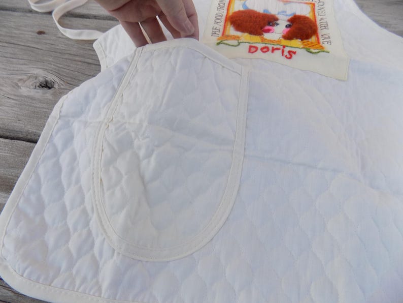 Op de afbeelding: Witte gewatteerde schort met een grote zak en een geborduurde patch met de tekst "The fool that thinks he can live on love alone" en "Doris".
