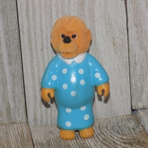May include: A vintage figurine of a bear character wearing a blue polka-dot pajama set with a white collar. The bear has orange fur and a black nose. The figurine is standing on a wooden surface.
