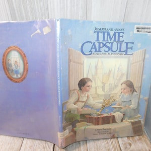 May include: A blue hardcover book titled "Joseph and Anna's Time Capsule: A Legacy from Old Jewish Prague" with an illustration of two children looking at a time capsule. The book cover features a light blue background with a white border.