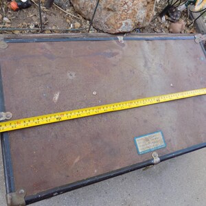 May include: An antique trunk with a brown exterior, black trim, and brass corner protectors. A yellow tape measure is laid across the trunk, showing a length of approximately 30 inches.