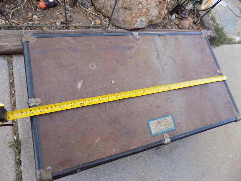 May include: An antique trunk with a brown exterior, black trim, and brass corner protectors. A yellow tape measure is laid across the trunk, showing a length of approximately 30 inches.