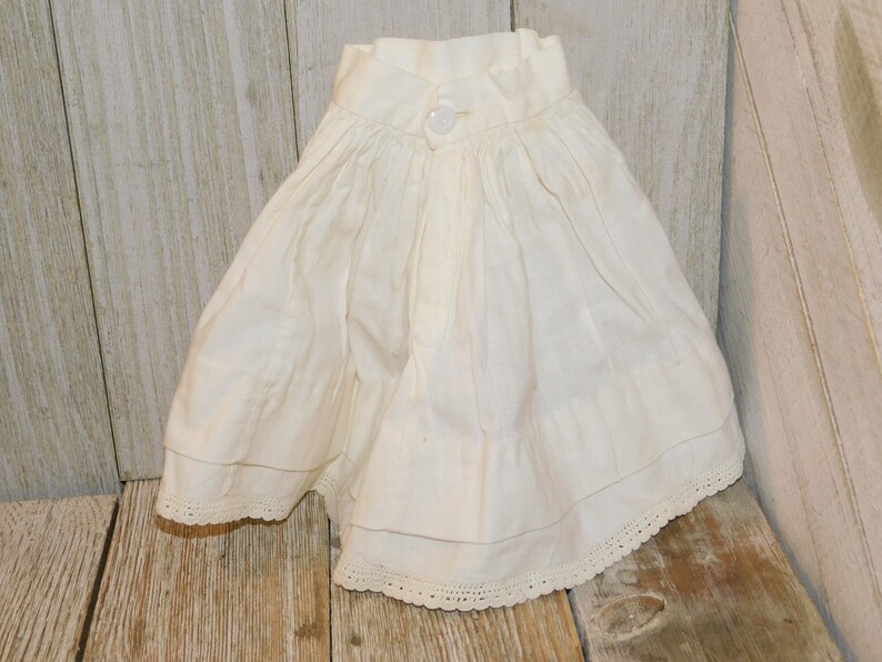 May include: A white cotton skirt with a button fastening and lace trim. The skirt is gathered at the waist and has a ruffled hem.