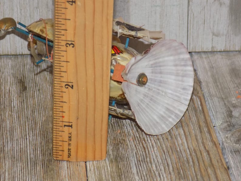 May include: A handmade art piece featuring a large white seashell with a button in the center. The artwork is constructed with various materials and is next to a wooden ruler, showing a measurement of approximately 3 inches.