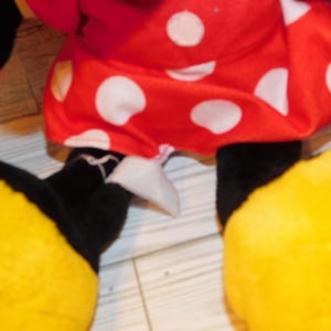 Op de afbeelding: Een pluche speelgoed met een rode jurk met witte stippen, zwarte benen, witte handschoenen en grote gele schoenen. Het speelgoed is waarschijnlijk een personage uit een populaire animatieserie.