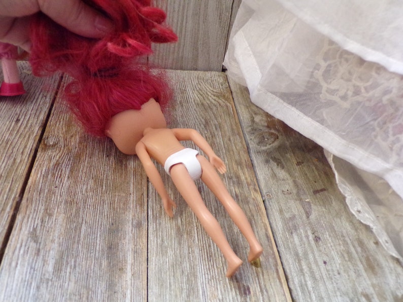 Pode incluir: Uma boneca com cabelo comprido e rosa brilhante deitada em uma superf&iacute;cie de madeira. A boneca est&aacute; sem cabe&ccedil;a e est&aacute; usando roupa interior branca.