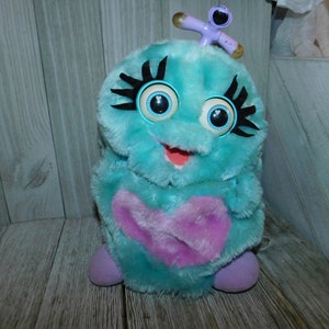 May include: A blue and purple plush toy with large eyes and a pink heart on its chest. The toy has a purple antenna with a small blue figure on top.