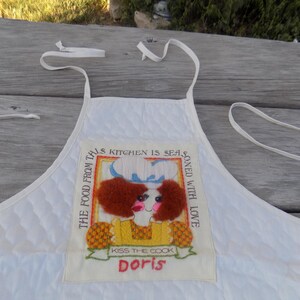 Op de afbeelding: Witte gewatteerde schort met een kleurrijk geborduurd patch waarop staat: "The food from this kitchen is seasoned with love. Kiss the cook Doris."