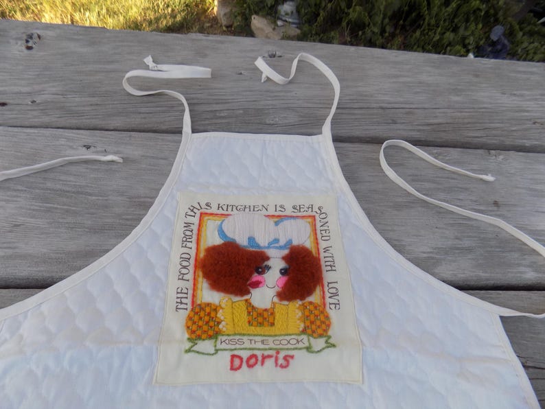 Op de afbeelding: Witte gewatteerde schort met een kleurrijk geborduurd patch waarop staat: "The food from this kitchen is seasoned with love. Kiss the cook Doris."