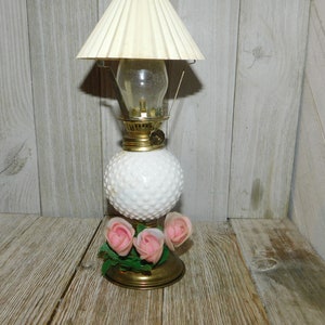 May include: A white milk glass oil lamp with a gold-toned base and a white lampshade. The lamp has a cluster of pink roses attached to the base.