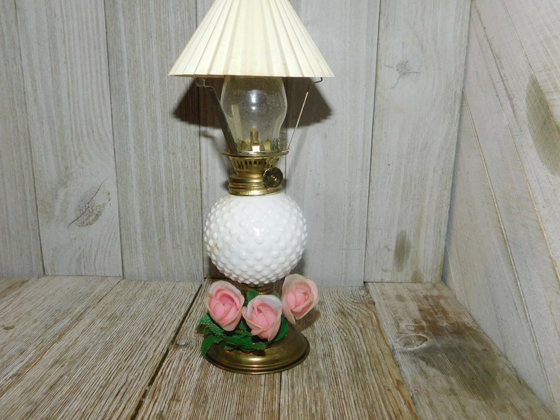 May include: A white milk glass oil lamp with a gold-toned base and a white lampshade. The lamp has a cluster of pink roses attached to the base.